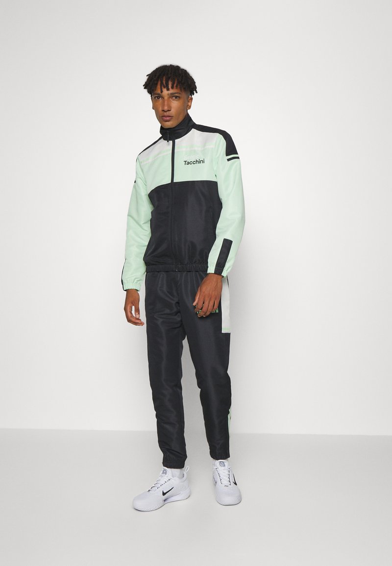 Träningsoverall i mintgrön och svart, med en zip-up jacka med kontrasterande paneler och matchande smalna byxor. Buren med vita sneakers.