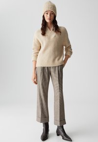 Maglione lavorato a maglia beige con scollatura ampia, abbinato a pantaloni grigi a quadri con risvolti. Il modello indossa un cappello di lana abbinato e stivali a punta.