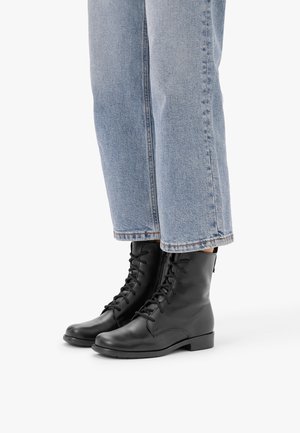 Person trägt hellblaue Jeans und schwarze Leder-Schnürstiefeletten und steht vor weißem Hintergrund.