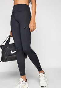 Μαύροι κολάν με ψηλή μέση και λεία υφή, με λευκό λογότυπο Nike, σε συνδυασμό με μαύρη αθλητική τσάντα και λευκά αθλητικά παπούτσια.