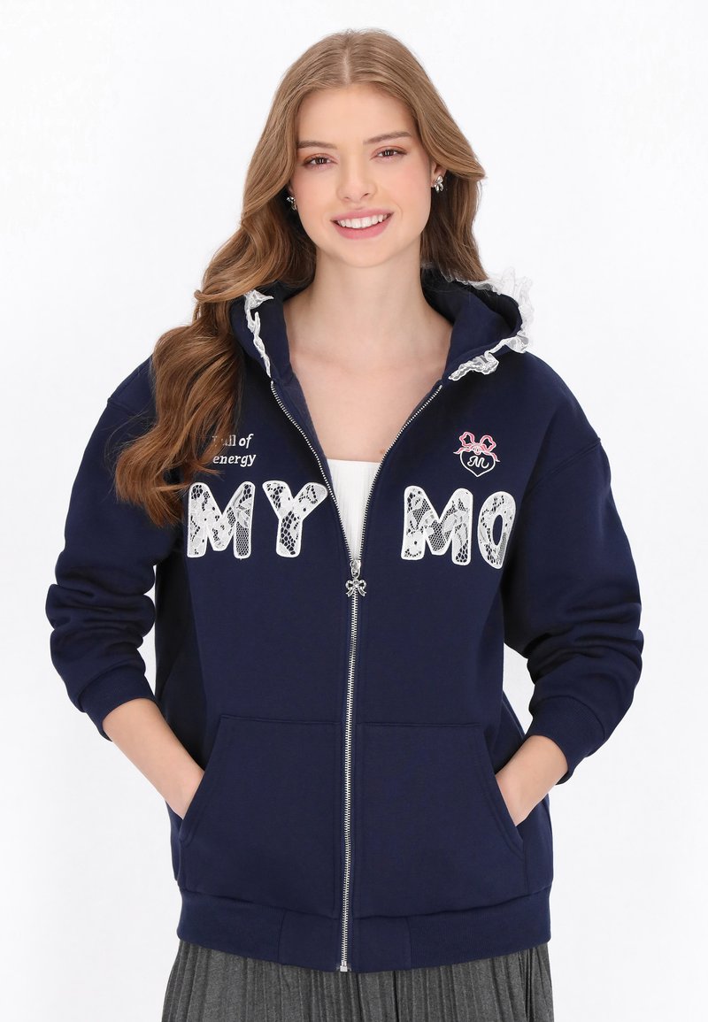 Sudadera con cremallera azul marino con ribete de encaje en la capucha, detalles grandes en letras blancas y bolsillos laterales. Incluye el texto: "lleno de energía" y "MY MO."