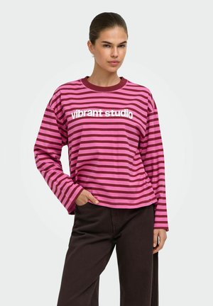 Femme aux cheveux attachés, portant un t-shirt à manches longues rayé rose et bordeaux avec le texte « vibrant studio » et un pantalon noir.