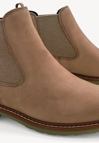 Bottines Chelsea en daim beige avec panneaux latéraux élastiques, coutures marron, languettes de tirage et semelles en caoutchouc vertes.