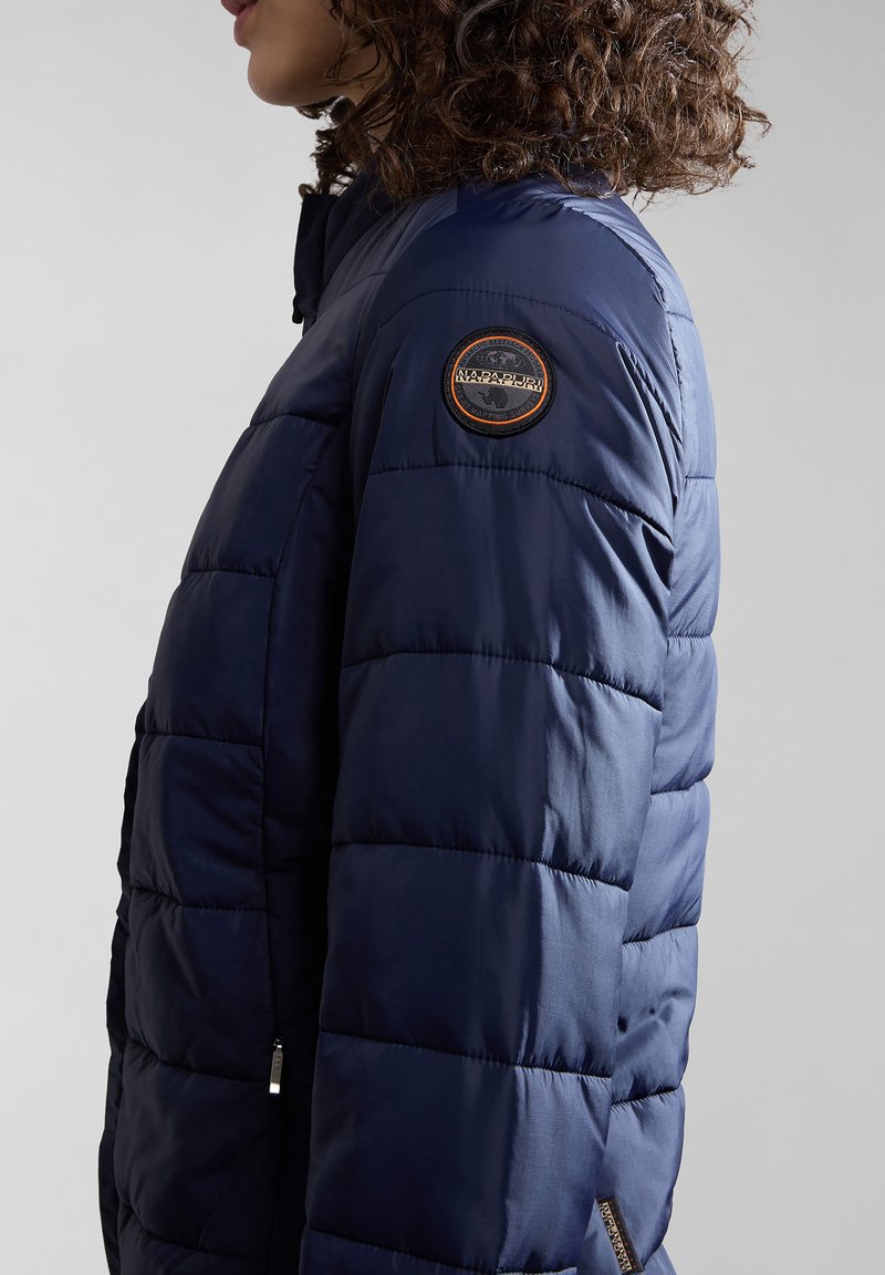 Giubbotto imbottito blu navy con design trapuntato, tasca con zip e una toppa con logo rotondo sulla spalla sinistra. Materiale morbido e leggero.