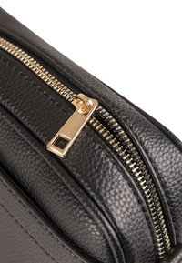 Coin de sac en cuir noir avec une fermeture éclair en métal doré et une surface texturée. La fermeture éclair est dotée d'une tirette rectangulaire et de dents visibles.