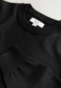 Pull noir à manches longues avec col rond et poignets côtelés, étiqueté "Next Regular Fit" en taille moyenne.