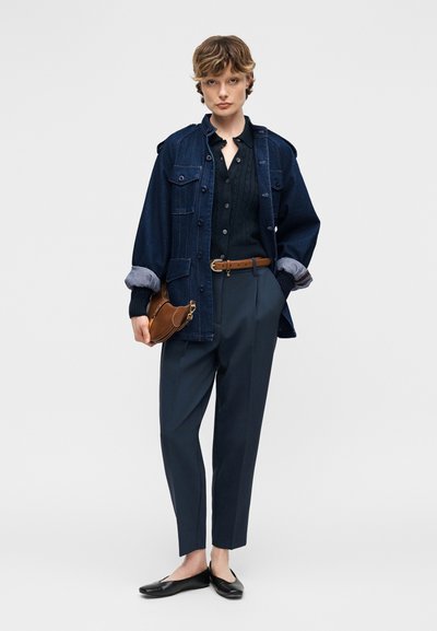 Ung person iført mørk denimjakke over sort knap-sweater, skræddersyede marineblå bukser, brunt bælte, holder brun håndtaske og sorte flade sko.