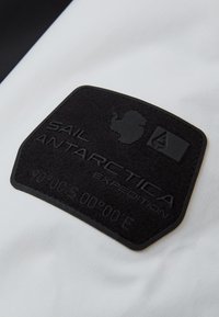 Svart texturerad lapp med präglad text "SAIL ANTARCTICA EXPEDITION," med en kartkontur av Antarktis, på vit tyg.