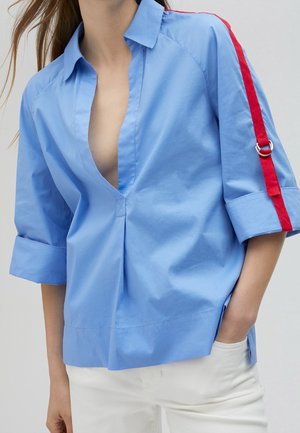 Vrouw die een losse lichtblauwe blouse draagt met een diepe V-hals en een rode streep op de bovenmouw, gecombineerd met een witte broek, hand in de zak.