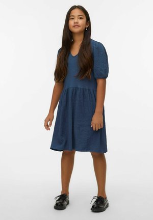 Niebieska teksturowana sukienka z dekoltem w kształcie litery V, krótkimi bufkami i marszczoną talią, zestawiona z czarnymi butami na sznurowadła.
