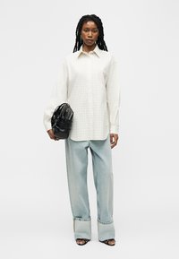 Γυναίκα με μακριά κοτσίδα, που φοράει φαρδιά λευκή πουκαμίσα, ανοιχτό μπλε τζιν παντελόνι με φάρδος, μαύρα σανδάλια και κρατάει μαύρη τσάντα.