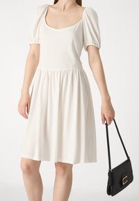 Robe blanche à manches bouffantes avec un décolleté carré et une taille froncée. Tissu lisse, jupe fluide, portée avec un sac à main noir avec un accent doré.