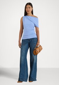 Kvinne iført en lyseblå asymmetrisk topp med én skulder, vide blå jeans, brune sko, holder en beige håndveske og har gullsmykker.