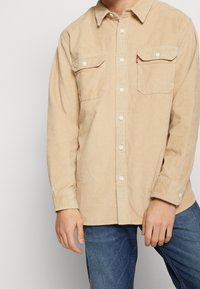 Beige corduroy overhemd met knopen, voorzien van twee borstzakken en witte knopen, met een puntige kraag en gestructureerde stof, gedragen met een blauwe spijkerbroek.