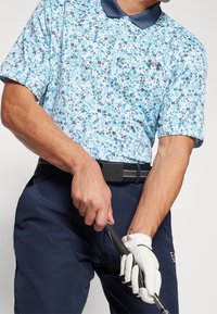 Polo bleu clair à motif floral avec manches courtes et col bleu marine. Porté avec un pantalon foncé et un gant de golf blanc, tenant un club de golf.