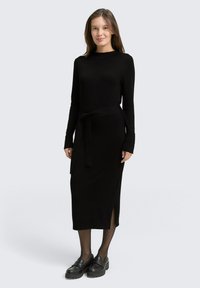 Schwarzes geripptes Strickkleid mit langen Ärmeln, einem taillierten Gürtel, einem seitlichen Schlitz und kombiniert mit schwarzen Plattformschuhen und transparenter Strumpfhose.