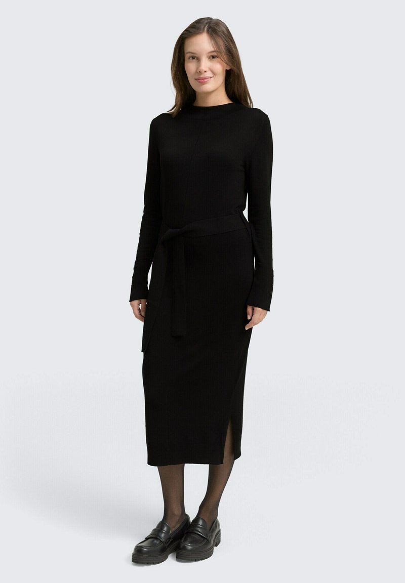 Schwarzes geripptes Strickkleid mit langen Ärmeln, einem taillierten Gürtel, einem seitlichen Schlitz und kombiniert mit schwarzen Plattformschuhen und transparenter Strumpfhose.