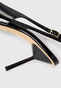 Scarpa slingback nera in pelle lucidante con tacco a stiletto sottile e fibbia dorata su una sottile cinghia alla caviglia, su sfondo bianco.
