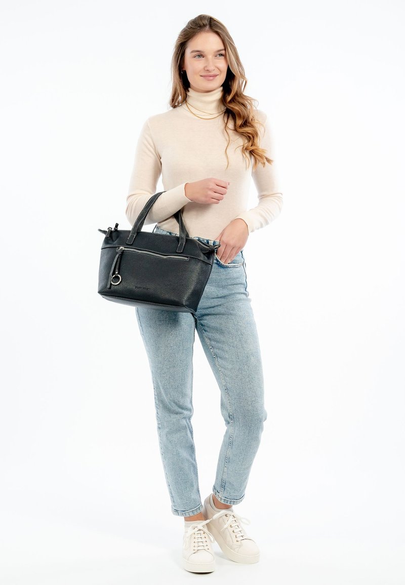 Svart läderhandväska med texturerad yta, dubbla handtag, silverfärgad detalj och zippered fack. Modellen bär ljusa jeans och en beige topp.