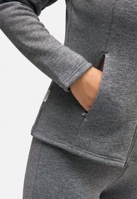 Chaqueta deportiva gris con una textura suave, que cuenta con un bolsillo con cremallera en el costado y un cuello tipo alto. La tela tiene un patrón jaspeado.
