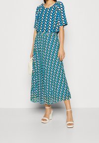 Blå og grøn mønstret kjole med en løs pasform, korte ærmer og en bæltestrop i taljen. Et par hvide kilehæle fuldender looket.