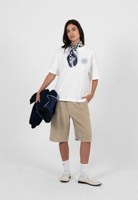 Femme portant un T-shirt blanc surdimensionné, un short beige jusqu'aux genoux, des mocassins blancs, des chaussettes blanches, un foulard à motifs autour du cou, tenant une veste marine dans la main gauche.