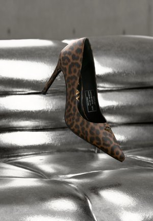Scarpa con tacco alto e punta a stampa leopardata poggiata su una superficie imbottita lucida e argento metallizzato.