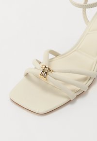 Sandale à talons beiges avec un bout carré, présentant de fines lanières croisées et une boucle décorative en ton doré. Texture lisse et design minimal.