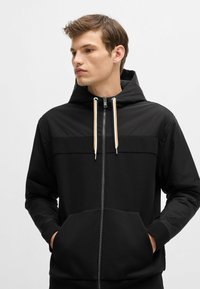 Felpa con zip nera dal design semplice, presenta un pannello superiore in contrasto, cappuccio regolabile con coulisse e tasche frontali. Texture liscia.