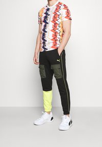 Pánský sportovní outfit s tričkem s multikolorovým zigzag vzorem a černými tepláky se zelenými kapsami a žlutými akcenty. Bílé tenisky.
