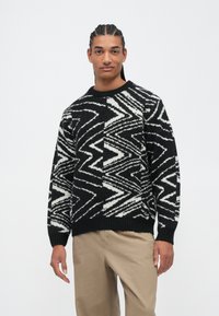Junger Mann mit geflochtenem Haar trägt einen schwarz-weißen Pullover mit Zickzackmuster und beigen Hosen, steht vor einem schlichten hellen Hintergrund.