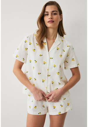 Jonge vrouw die een witte korte mouw blouse en bijpassende korte broek met kleine gele citroenprints draagt, staand tegen een effen achtergrond.