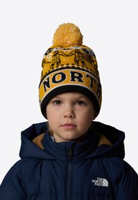 Żółta wełniana czapka z czarnymi akcentami, z białym pomponem i graficznym wzorem. Wyraz "NORTH" jest wyraźnie eksponowany.