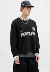 Fekete, hosszú ujjú pulóver bordás textúrával, kerek nyakkal, fehér részletekkel és grafikával, amelyen a "VENTURA" felirat szerepel. Sötét cargo rövidnadrággal párosítva.