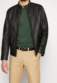 Černá kožená bunda s stojáčkovým límcem, zapínáním na zip a žebrovanými detaily na ramenou, přes zelenou košili a béžové kalhoty.