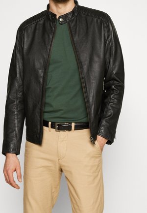 Zwarte leren jas met een staande kraag, ritssluiting en geribbelde details op de schouders, gedragen over een groene shirt en beige broek.