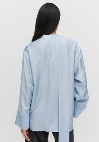 Vaaleansininen satiininen pitkähiha-paitapusero, jossa on korkea kaula-aukko ja takana solmittava yksityiskohta. Ominaisuuksiin kuuluvat leveät hihansuut ja rento istuvuus.