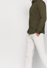 Olivgrünes Hemd zum Knöpfen mit einer einzelnen Brusttasche, kombiniert mit weißen Hosen. Der Stoff wirkt glatt mit sichtbaren Nähten und schwarzen Knöpfen.