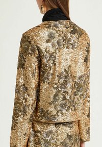 Veste en sequins dorés avec motif floral et col noir. Surface texturée reflétant la lumière, mettant en valeur un mélange de teintes dorées et brunes.