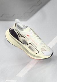 Nowoczesne beżowe i białe sneakersy z czarnymi i szarymi akcentami, marka "Stella McCartney", z grubą podeszwą, pokazane z bocznego widoku pod kątem.