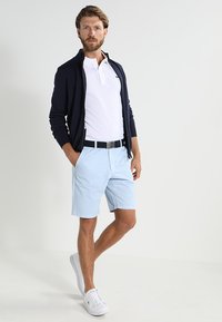 Cardigan blu navy con zip sopra una polo bianca, pantaloni corti azzurri, cintura nera, sneakers bianche. Maglietta a maniche corte, outfit estivo informale.