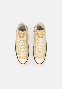 Tenisky vysokého střihu s žlutým květinovým vzorem, bílými tkaničkami, černou gumovou podrážkou a lesklou špicí. Hladký plátěný povrch.