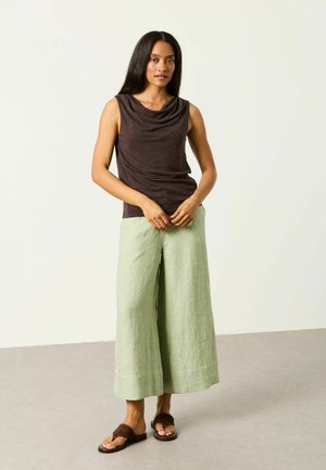 Frau steht und trägt ein ärmelloses dunkelbraunes Oberteil, hellgrüne Hosen mit weitem Bein und dunkelbraune Sandalen vor einem schlichten Hintergrund.