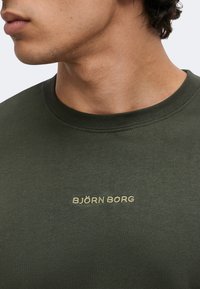 Gros plan sur le cou et la mâchoire d’une personne portant une chemise vert olive avec "BJÖRN BORG" brodé en fil clair sur la poitrine.