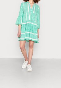 Vestido verde con estampado, escote en V, mangas acampanadas y diseño en capas. Presenta rayas y motivos geométricos. Llevado con zapatillas blancas.