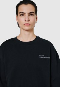 Svart sweatshirt med rund halsringning, prydd med vit broderad text "HALO 19681972198" på vänster sida. Mjuk tygkvalitet och avslappnad passform.
