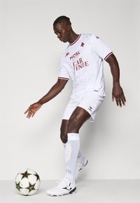 Weißes Fußballtrikot mit roten Akzenten und grafischen Mustern, kombiniert mit weißen Shorts und Socken sowie schwarz-weißen Fußballschuhen. Ein Ball in der Nähe.