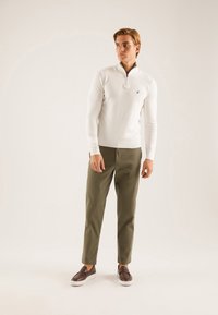 Maglione bianco a mezza zip, attillato, con polsini e orlo a costine. Pantaloni verde oliva, a taglio dritto, con scarpe brown senza lacci. Logo discreto sul maglione.