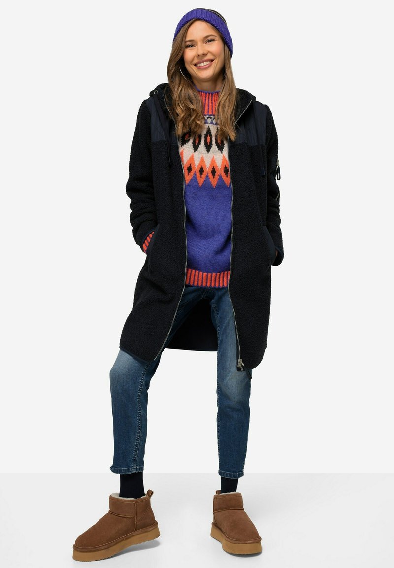 Cappotto in pile nero con zip, maglione blu con motivi geometrici, jeans e stivali alla caviglia marroni. Il modello indossa un berretto di maglia viola.
