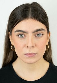 Una donna con lunghi capelli scuri e pelle chiara, che indossa piccoli orecchini a cerchio dorati e una maglietta nera, fissa direttamente lo spettatore.